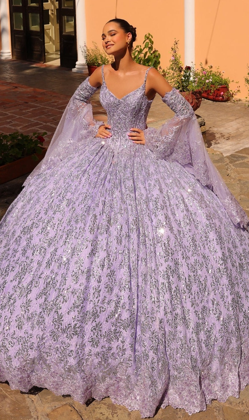 Smiling with her eyes closed, a woman stands outdoors wearing the Quinceanera Dress 54303 by Amarra, featuring lavender fabric with intricate silver patterns and off-shoulder sleeves.